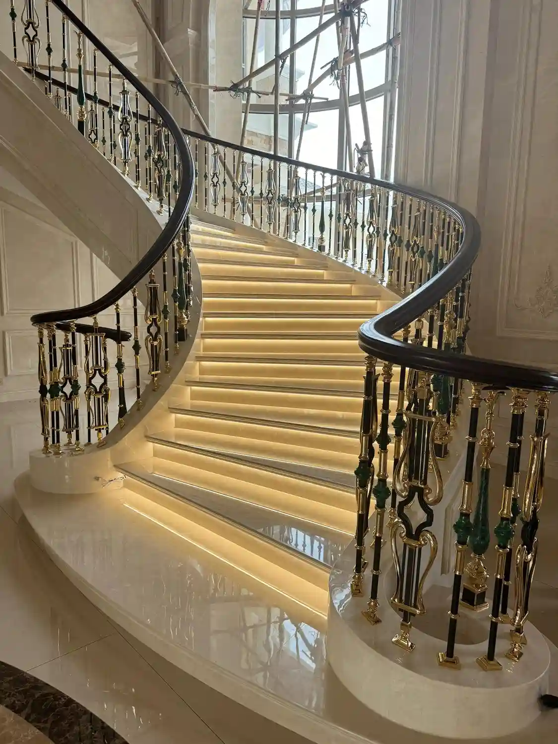 Dubbel gebogen trap van keizerlijk marmer met crèmekleurige marmeren treden, geïntegreerde warme LED-trapverlichting, sierlijke balustrade van messing en donkergroen, donker gepolijste stenen leuning, neoklassiek gebogen trapontwerp in de lobby van een luxe hotel of paleis