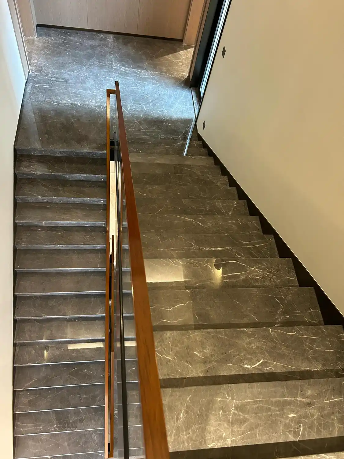 Moderne donkergrijze marmeren trap met vrijdragende, zwevende treden en een glazen balustrade, gepolijste marmeren treden en stootborden in Pietra Grey of Armani Grey met opvallende witte aders, een leuning van walnotenhout, een eigentijds, luxueus interieurontwerp met marmeren trapbekleding