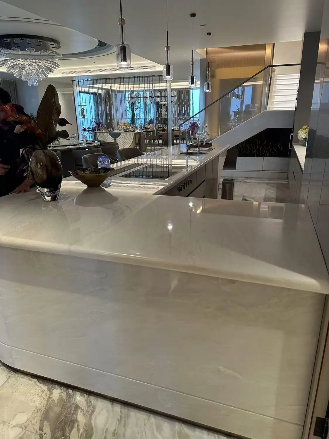 Groot, gebogen keukeneiland en barblad in honingkleurig onyx of doorschijnend crèmekleurig marmer, spiegelglansafwerking met warme gouden aders, watervalrand, geïntegreerde kookplaat, platen in boekpatroon die een sculpturaal middelpunt vormen in een luxe, eigentijdse keuken