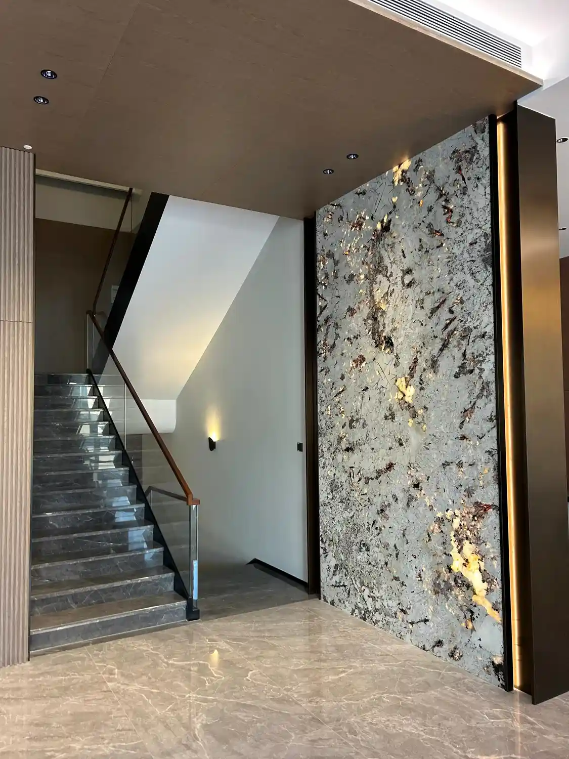 Contemporary luxury entryway featuring a floor-to-ceiling backlit translucent stone wall panel with warm honey-amber glow revealing copper, bronze, and burgundy mineral inclusions in a chaotic organic pattern, framed by dark bronze metal trim with concealed edge LED strip lighting, paired with cantilevered floating gray marble staircase treads with white veining and frameless tempered glass balustrade with walnut handrail, vertical warm-medium-brown oak wood slat ceiling with recessed downlights, large-format polished beige porcelain floor tiles, modern minimalist gallery aesthetic combining organic natural stone artistry with industrial precision craftsmanship