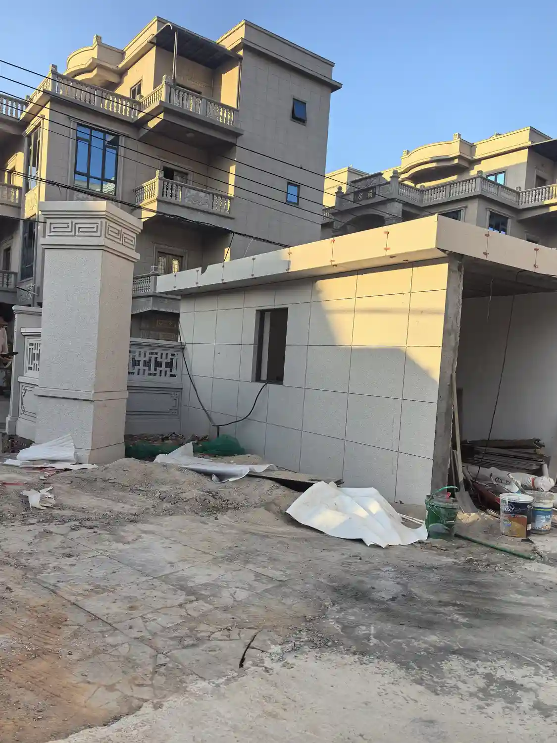 Chinese luxury villa exterior during final construction phase featuring decorative white rendered stone gate pillars with carved classical Chinese huiwen geometric meander key pattern relief and stepped molded capitals, spindle stone balusters on upper balconies, carved stone panels beneath balcony railings, arched entryway with carved Chinese calligraphy reading Lanzhi, white ceramic tile cladding with glossy reflective finish on ancillary small building, warm golden-hour afternoon sunlight illuminating cream stucco and beige facades, construction materials including white tarps, paint buckets, and green garden hose scattered in courtyard, traditional Chinese ornamental motifs blended with Western villa architectural forms