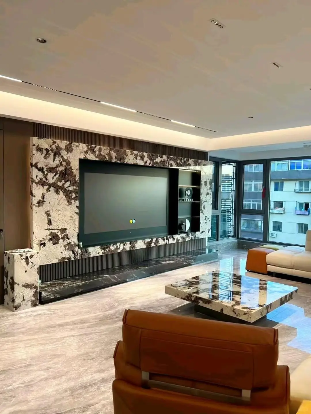 Ultra-modern masculine luxury penthouse living room centered on a dramatic exotic brecciated Italian marble TV wall resembling Patagonia or Breccia Capraia with warm cream-ivory base and bold chocolate brown, black, and rust-brown fragmented veining, bookmatched slabs forming wraparound feature wall with cantilevered shelf above and flanking side panel, polished Nero Marquina black marble hearth strip with subtle white veining along floor base, matching exotic marble rectangular coffee table with beveled polished edges, large-format polished light gray marble floor tiles, dark walnut vertically fluted wood panels adding texture, caramel leather lounge chair with matching ottoman, cream modern modular sectional sofa, floor-to-ceiling windows with dark aluminum frames showing urban high-rise view