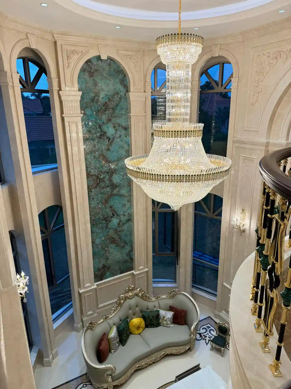 Opulent palatial double-height grand hall photographed from upper landing overlooking the room below, centered on towering backlit translucent green onyx feature wall panel spanning full two-story height between three grand Palladian arched windows with dark wooden mullions revealing deep blue twilight sky, soft mint sea-green base color interlaced with white, cream, and warm amber-brown veining glowing from concealed LED backlighting, framed by classical cream-painted pilasters and decorative cornices, ornate carved and gilded gold-leaf French Louis XV-style settee with sage green velvet upholstery and decorative pillows in mustard, burgundy, and patterned silks, spectacular multi-tier crystal chandelier with cascading strands, polished cream marble floor with decorative inlay border, black-and-white geometric marquetry stone floor inlay framing seating area in Greek-key scroll motif