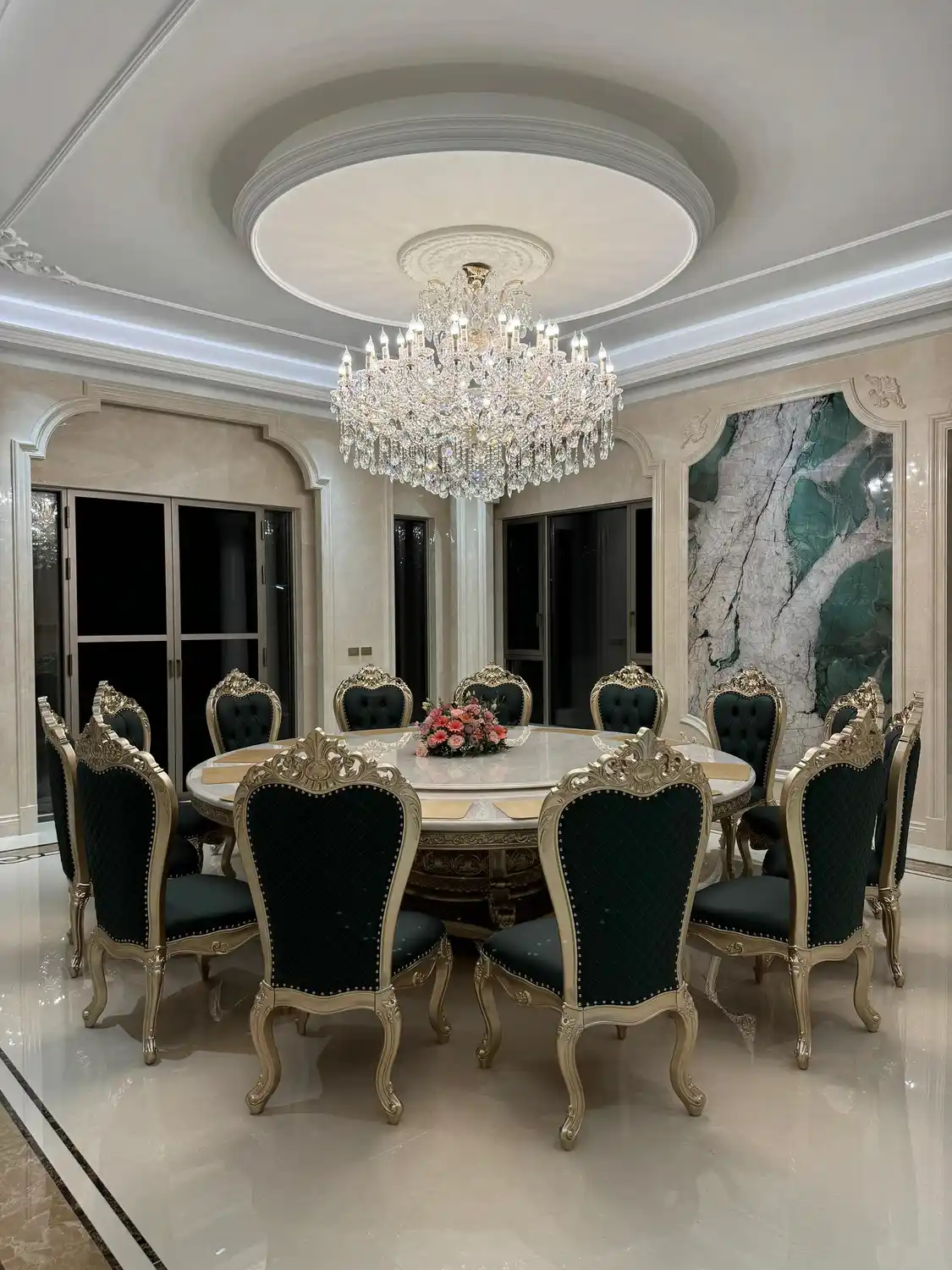 Opulent Baroque-Rococo formal dining room centered on dramatic large-scale bookmatched decorative wall panel of exotic green marble or Calacatta Verde with jade-emerald green veining accented by rose pink, gold, and gray mineral inclusions on white-cream base, mirror-symmetric installation creating butterfly-like composition, high-gloss polished finish reflecting grand multi-tier crystal chandelier with cascading hand-cut crystal pendants, framed by classical white molding with subtle carved corner ornaments, mirrored back wall behind ten-seat round dining table expanding spatial perception, polished white Carrara or Calacatta marble tabletop with central rotating lazy Susan, ten ornate Louis XV-style chairs with carved champagne-gold leaf frames and tufted emerald-green velvet upholstery with brass nailhead trim, polished cream beige marble flooring with dark stone border inlay, tray ceiling with oval medallion