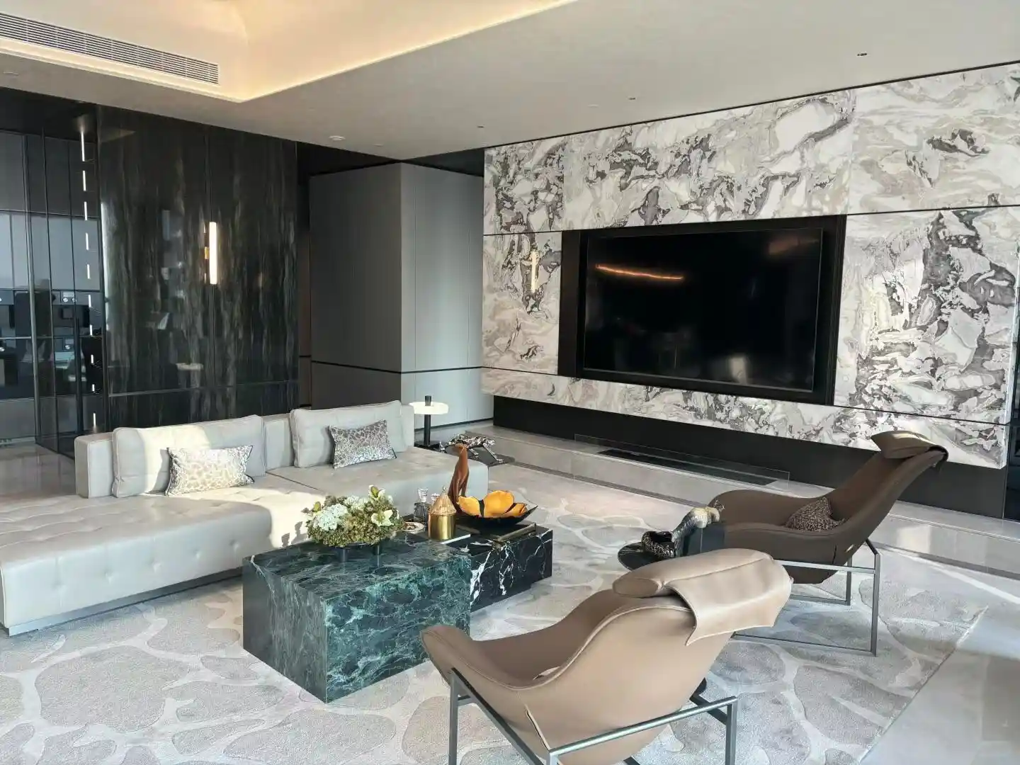 Ultra-modern contemporary luxury living room featuring floor-to-ceiling bookmatched Calacatta or Arabescato-style Italian marble feature wall behind large flat-screen television, warm cream-ivory base with dramatic bold gray-charcoal and warm golden veining mirrored across central seam creating symmetrical butterfly pattern, highly polished mirror finish reflecting cove and recessed ceiling lighting, framed by thin dark metal trim lines, adjacent dark bookmatched black onyx or marble side panels with subtle vertical veining, two monolithic rectangular Verde Alpi or Verde Guatemala deep emerald-green marble coffee tables with white and lighter-green veining, large L-shaped light gray sectional sofa with deep button-tufted capitone base detailing, two cream leather cocoon-style modern Italian lounge chairs with chrome sled bases, glass-fronted display cabinet, sculptural gold brass decorative objects and fresh white hydrangea floral arrangement on coffee table