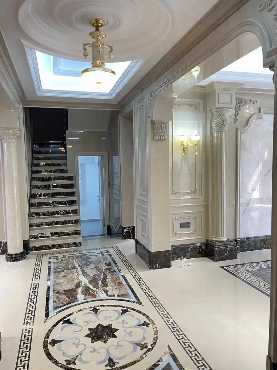 Neoclassical European luxury foyer featuring Crema Marfil marble flooring with intricate waterjet-cut Greek key border patterns and floral medallion inlay, complemented by Emperador Dark marble staircase with classical carved balusters and risers, ornate carved marble columns framing arched entryways, high-gloss polished finish reflecting crystal chandelier light, neoclassical European interior design combining Mediterranean villa grandeur with artisan stone craftsmanship for upscale residential and hospitality foyer installations