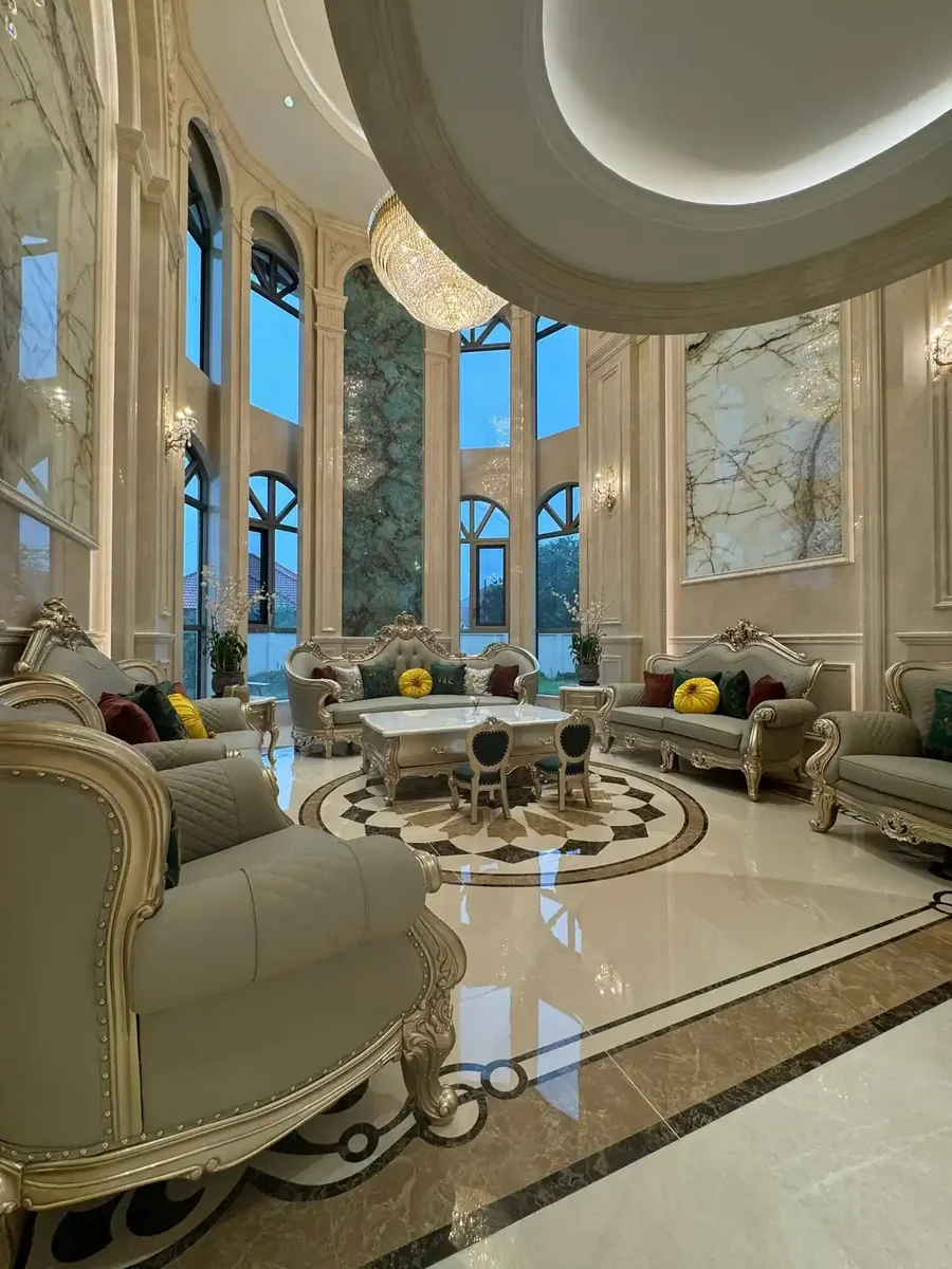 Opulent double-height European neoclassical living room clad in Calacatta Gold marble wall panels featuring warm cream white ground with dramatic gold veining throughout, complemented by Verde Guatemala green marble accents and geometric inlaid marble flooring medallion, neoclassical baroque luxury living room with crystal chandelier and classical plaster moldings, high-gloss polished marble surface reflecting ambient lighting, premium luxury residential interior combining Calacatta gold's theatrical veining with verde Guatemala's exotic green for palatial open-plan living spaces