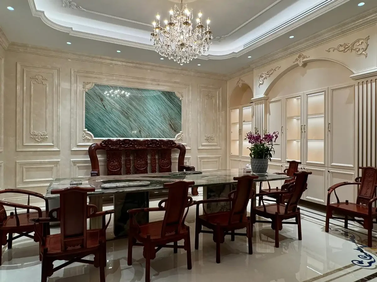 Luxurious neoclassical European dining room featuring Rainforest Green marble accent wall with dramatic flowing diagonal veining resembling forest canopy, complemented by a polished Rainforest Green marble tabletop, ornate carved wood moldings and crystal chandelier overhead, classical European paneling framing the marble feature wall, polished finish enhancing the deep emerald green coloration, premium luxury residential dining space combining exotic brecciated marble with neoclassical architectural details for high-end private dining rooms and formal entertaining areas