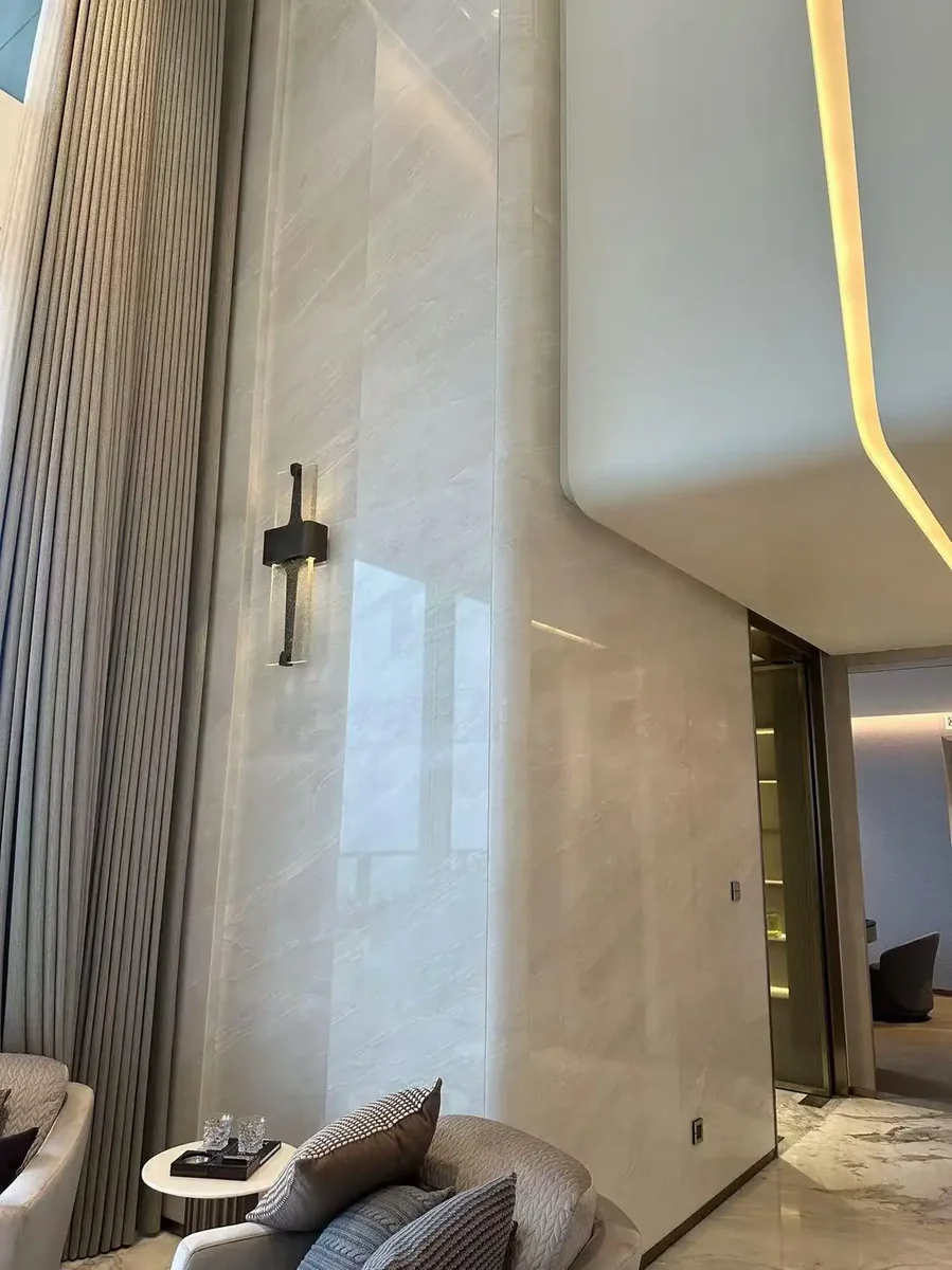 Tall double-height contemporary luxury living room featuring Botticino cream beige marble wall cladding with subtle linear horizontal veining, modern brass wall sconce providing ambient illumination, floor-to-ceiling neutral drapery, curved upholstered sofa, and elegant recessed ceiling lighting creating a sophisticated minimalist atmosphere, polished marble surface reflecting warm tones, premium luxury residential modern living space combining Italian Botticino marble's understated elegance with contemporary interior architecture for refined open-plan living areas