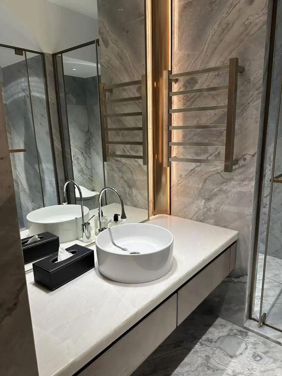 Luxurious contemporary bathroom clad in Blue Roma marble wall cladding featuring blue-grey ground with dramatic flowing white veining, complemented by white marble vanity countertop and undermount sink, gold-toned heated towel rails, modern chrome fixtures, and concealed ambient lighting creating a sophisticated spa atmosphere, polished marble finish enhancing depth of color, premium luxury residential bathroom combining exotic Blue Roma marble's dramatic character with contemporary spa design for upscale master bath retreats