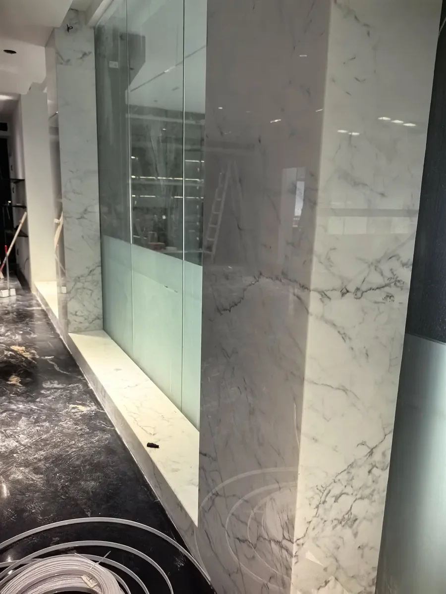Luxurious contemporary commercial lobby clad in white Carrara marble wall cladding featuring classic grey veining throughout, complemented by Carrara marble bench seating with highly polished reflective surfaces, modern recessed lighting and clean architectural lines creating an upscale corporate or hotel aesthetic, premium quality Italian marble installation for commercial reception areas and corporate lobbies, combining timeless Carrara marble's classical beauty with modern minimalist commercial interior design for sophisticated public spaces